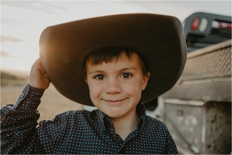 young cowboy smiles at camera with a hand on his hat at sunset
