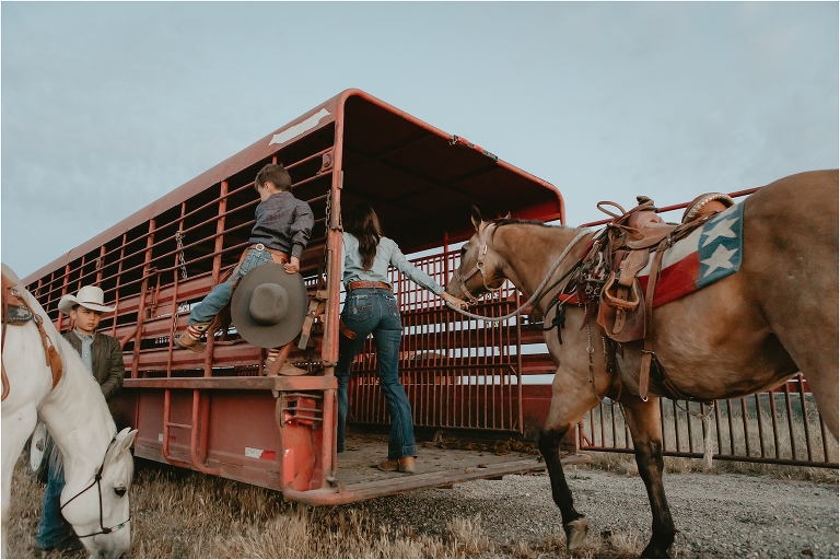 cowgirl loads buckskin horse into horse trailer while cowboy son watches