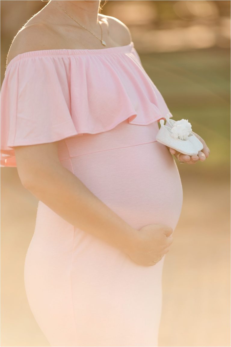 Pregnant woman in pink dress holding baby shoes by Elizabeth Hay Photography