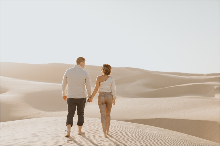 couple holding  hands and walking on beach dunes in Pismo Beach California by Elizabeth Hay Photography
