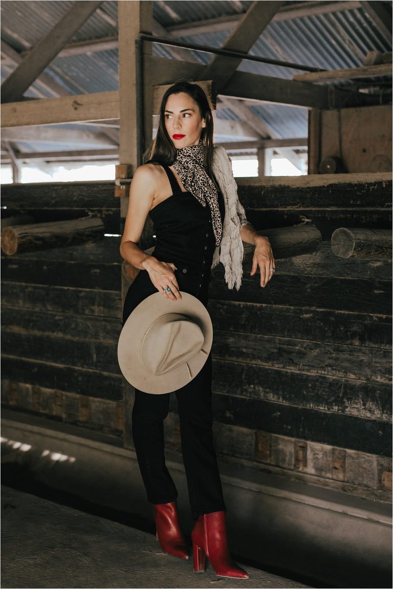 Western Fashion Inspo with Lindsay Branquinho in barn aisle wearing black overalls, a vintage Stetson hat,  cheetah wild rag and red lipstick and booties by Elizabeth Hay Photography.