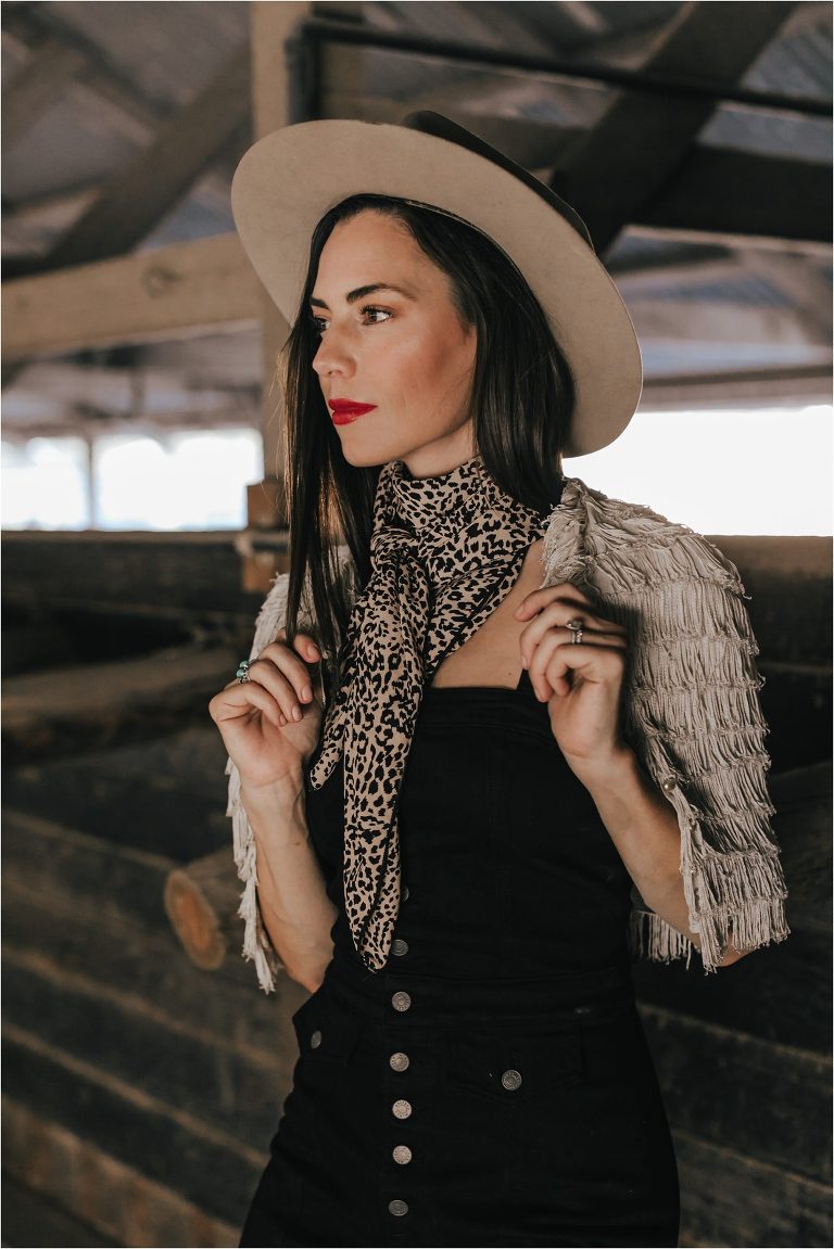 Western Fashion Inspo with Lindsay Branquinho in barn aisle wearing black overalls, a vintage Stetson hat,  cheetah wild rag and red lipstick and booties and a fringe shawl by Elizabeth Hay Photography.