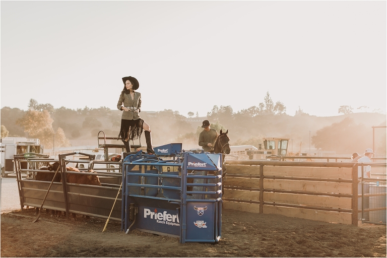 Lindsay Branquinho standing on top of a Priefert Ranch Equipment roping chute wearing a black Stetson hat, fringe skirt and tweed blazer by California Equine Photographer Elizabeth Hay Photography. 